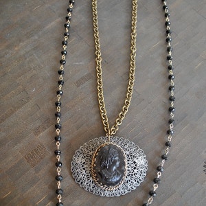 May include: A gold chain necklace with a black cameo pendant and a black beaded chain. The cameo features a woman's profile.