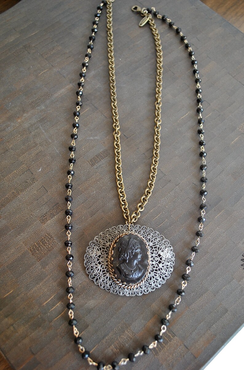 May include: A gold chain necklace with a black cameo pendant and a black beaded chain. The cameo features a woman's profile.