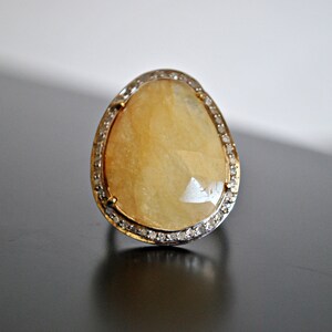 May include: A large, teardrop-shaped ring with a yellow gemstone. The gemstone is surrounded by a band of small, clear stones. The ring's band is gold-colored. The ring is set against a dark surface.