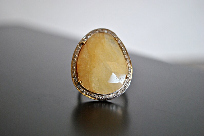 May include: A large, teardrop-shaped ring with a yellow gemstone. The gemstone is surrounded by a band of small, clear stones. The ring's band is gold-colored. The ring is set against a dark surface.