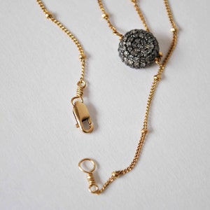 May include: A gold-colored necklace with a round, dark gray, gem-studded pendant. The necklace has a delicate chain with small gold beads and a clasp. The pendant is the focal point, adding a touch of sparkle and sophistication.