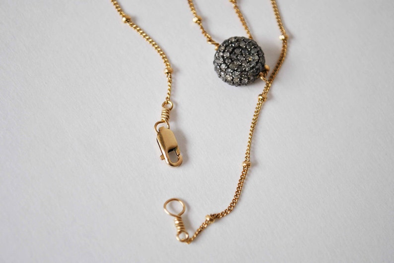 May include: A gold-colored necklace with a round, dark gray, gem-studded pendant. The necklace has a delicate chain with small gold beads and a clasp. The pendant is the focal point, adding a touch of sparkle and sophistication.