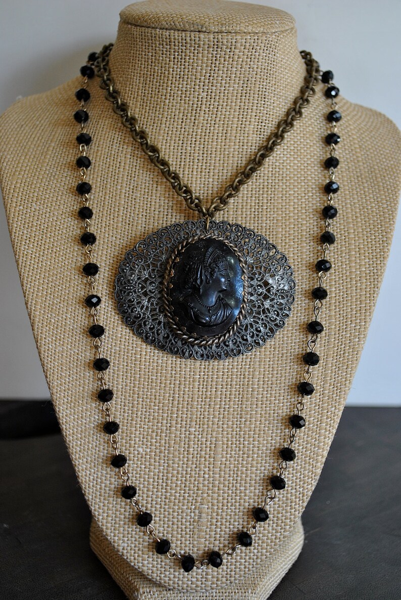 May include: A black cameo pendant necklace with a filigree design and a black beaded chain. The cameo features a woman's profile.