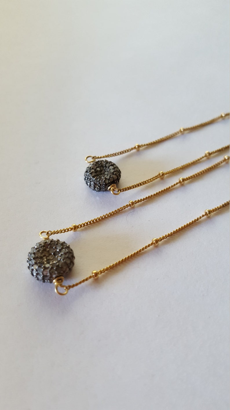 May include: Gold-toned necklaces with a delicate chain design. Each necklace features a small, round, textured pendant with a dark, sparkling surface. The necklaces are displayed on a white background.