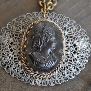 May include: A black cameo pendant featuring a woman's profile, set in a gold-toned filigree frame. The pendant is attached to a chain with black beads.