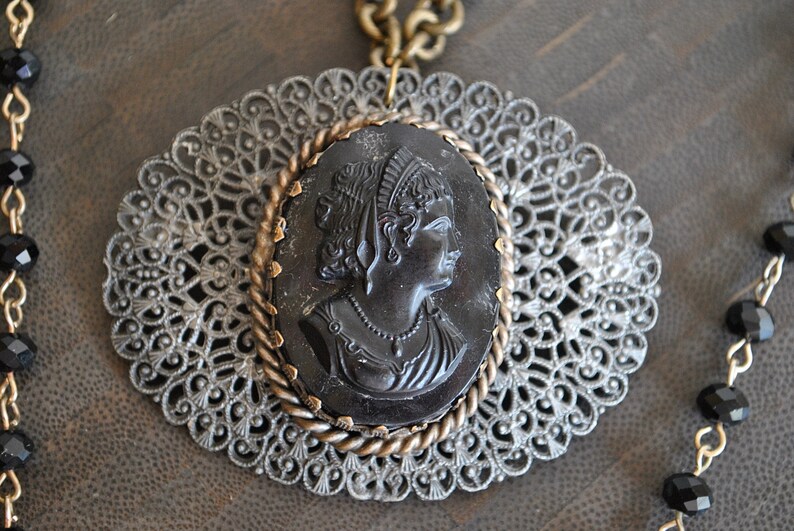 May include: A black cameo pendant featuring a woman's profile, set in a gold-toned filigree frame. The pendant is attached to a chain with black beads.