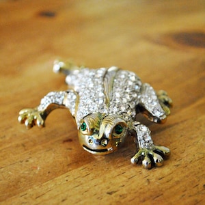May include: A silver and white jeweled frog brooch with green eyes. The frog is sitting on a wooden surface.