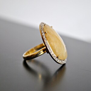 May include: A gold ring with a large, oval, yellow gemstone. The gemstone is surrounded by a row of small, clear stones. The ring is set on a dark surface.