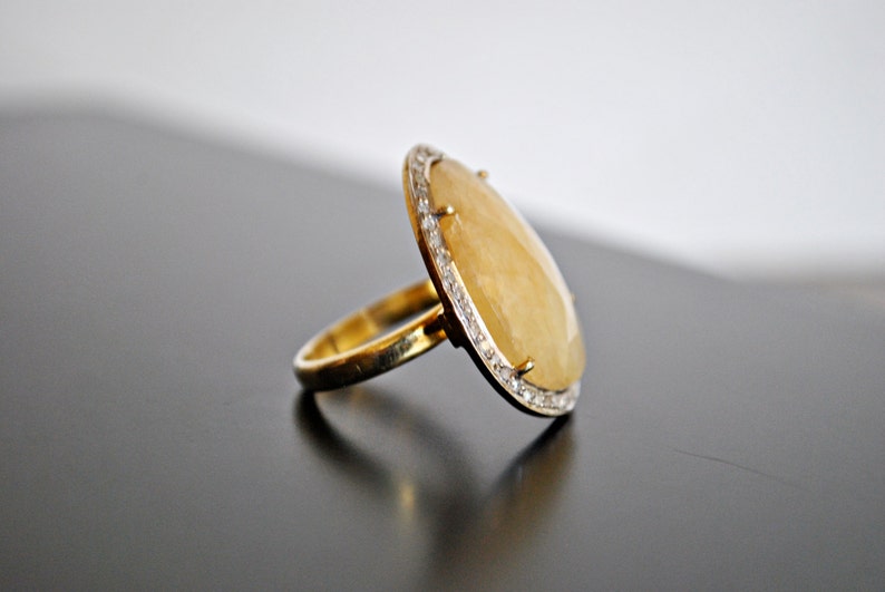May include: A gold ring with a large, oval, yellow gemstone. The gemstone is surrounded by a row of small, clear stones. The ring is set on a dark surface.