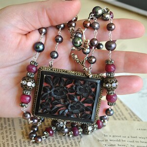 May include: A vintage necklace featuring a large, rectangular pendant with a carved floral design. The necklace incorporates dark gray pearls, red gemstones, and ornate metal accents. The pendant is black with a red background.