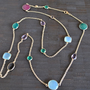 May include: A gold chain necklace with a variety of gemstone pendants, including green, blue, purple, and pink. The necklace is on a dark wooden surface.