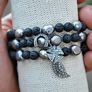 May include: Three layered bracelets with black lava rock beads and silver charms. The charms are a horseshoe, a cross, and a feather. The bracelets are on a white fabric background.
