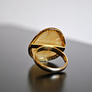 May include: A gold ring with a large, light-colored, oval-shaped stone. The ring's band and setting are gold. The stone has a pearlescent appearance. The ring is set against a dark surface.