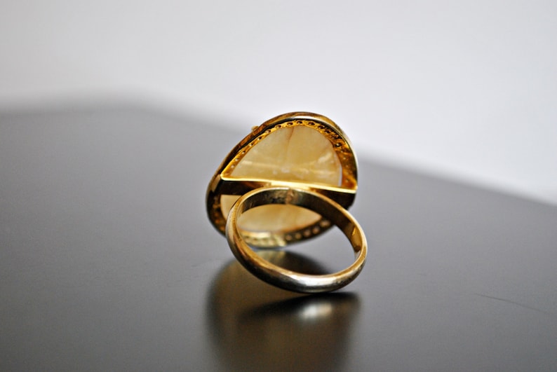 May include: A gold ring with a large, light-colored, oval-shaped stone. The ring's band and setting are gold. The stone has a pearlescent appearance. The ring is set against a dark surface.