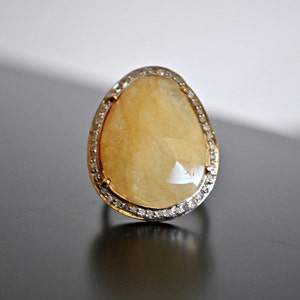 May include: A large, teardrop-shaped ring with a faceted, pale yellow gemstone. The stone is set in a gold band, surrounded by a halo of small, clear stones. The ring is displayed on a dark surface.