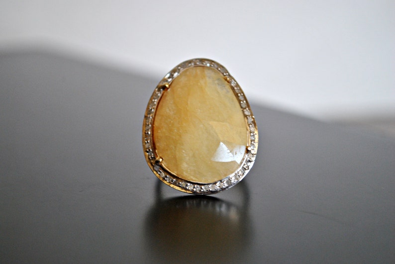 May include: A large, teardrop-shaped ring with a faceted, pale yellow gemstone. The stone is set in a gold band, surrounded by a halo of small, clear stones. The ring is displayed on a dark surface.