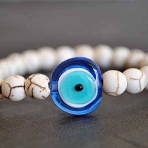 May include: White beaded bracelet with a blue evil eye charm. The charm has a white center with a blue circle and a black pupil.