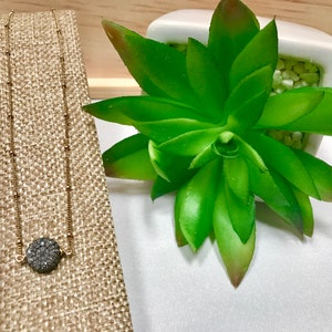 May include: A gold-colored necklace with a round, dark gray druzy pendant. The necklace is displayed on a tan fabric stand next to a green succulent plant in a white pot. The necklace chain has small, evenly spaced beads.