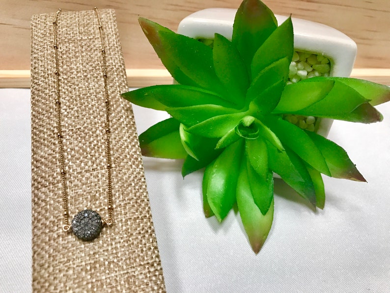 May include: A gold-colored necklace with a round, dark gray druzy pendant. The necklace is displayed on a tan fabric stand next to a green succulent plant in a white pot. The necklace chain has small, evenly spaced beads.