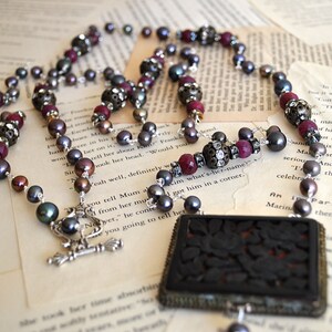 May include: A necklace with dark gray pearls, red beads, and ornate silver accents. The necklace features a rectangular pendant with a carved floral design. The jewelry rests on vintage book pages.