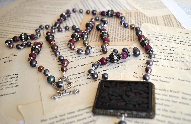 May include: A necklace with dark gray pearls, red beads, and ornate silver accents. The necklace features a rectangular pendant with a carved floral design. The jewelry rests on vintage book pages.