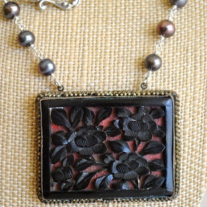 May include: A necklace featuring a rectangular pendant with carved black floral design on a red background. The pendant is framed in a gold-toned metal. The necklace has a silver chain with dark gray and brown pearls.