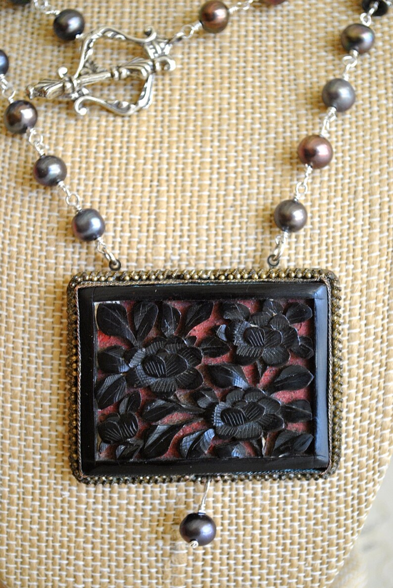 May include: A necklace featuring a rectangular pendant with carved black floral design on a red background. The pendant is framed in a gold-toned metal. The necklace has a silver chain with dark gray and brown pearls.