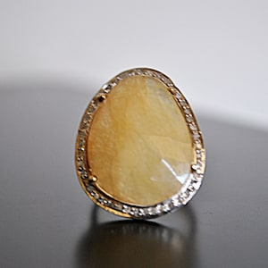May include: A pear-shaped ring with a large, light yellow gemstone. The gemstone is surrounded by a silver-colored band with small, clear stones. The ring is set on a dark surface.