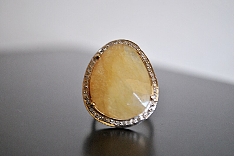 May include: A pear-shaped ring with a large, light yellow gemstone. The gemstone is surrounded by a silver-colored band with small, clear stones. The ring is set on a dark surface.
