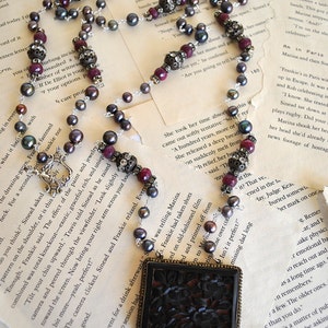 May include: Elegant necklace featuring dark gray pearls, ruby-colored beads, and ornate silver accents. The focal point is a rectangular pendant with a carved floral design, creating a vintage aesthetic.