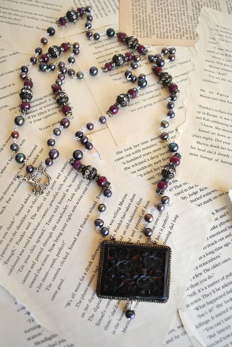 May include: Elegant necklace featuring dark gray pearls, ruby-colored beads, and ornate silver accents. The focal point is a rectangular pendant with a carved floral design, creating a vintage aesthetic.