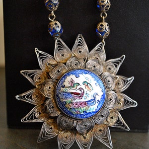 May include: A silver filigree necklace with a blue and white enamel pendant featuring a floral design and birds. The pendant is surrounded by a silver starburst.