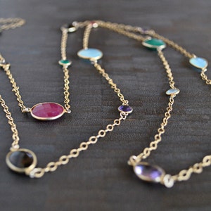 May include: Two gold-toned necklaces with colorful gemstones. The necklaces feature a chain design with oval and round gemstones in shades of red, green, blue, purple, and brown. The necklaces are displayed on a dark wooden surface.