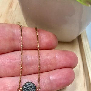 May include: A delicate gold necklace featuring a round, dark gray pave diamond pendant. The pendant is suspended from a fine gold chain with small, evenly spaced gold beads. The necklace is displayed against a neutral background.