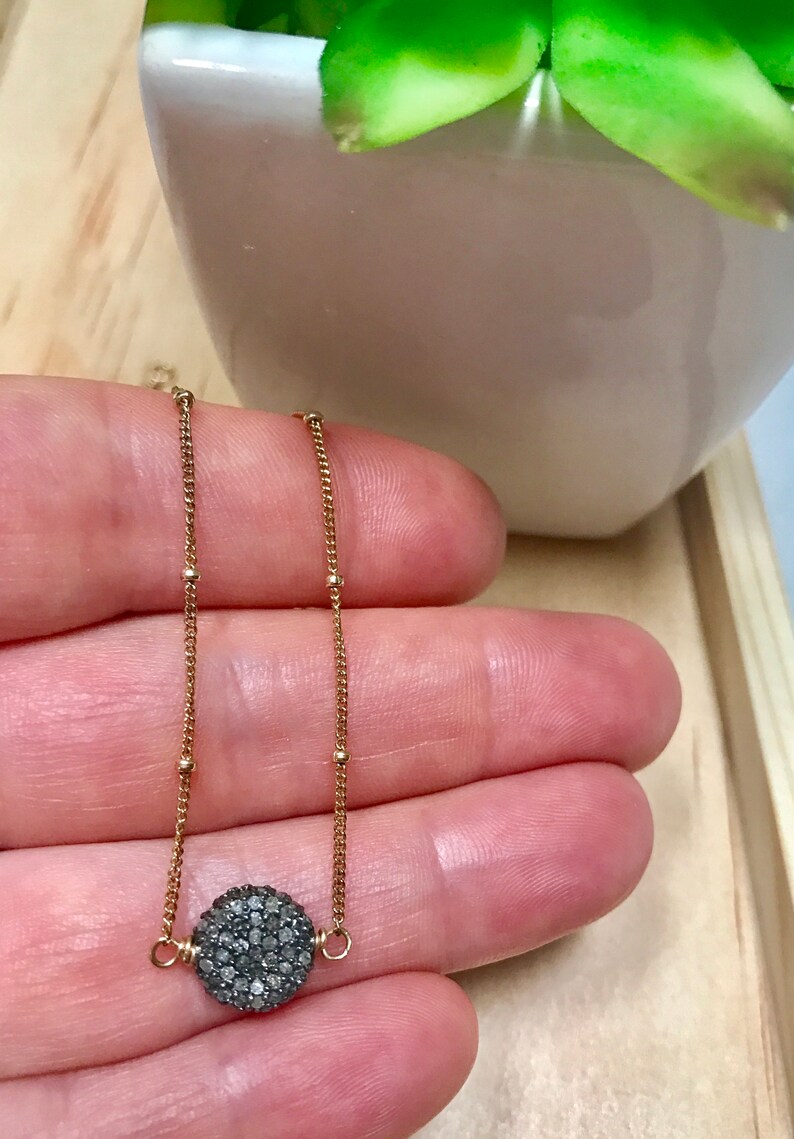 May include: A delicate gold necklace featuring a round, dark gray pave diamond pendant. The pendant is suspended from a fine gold chain with small, evenly spaced gold beads. The necklace is displayed against a neutral background.