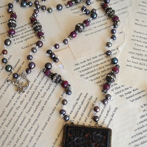 May include: A long necklace featuring dark gray pearls, burgundy beads, and ornate silver accents. The necklace has a rectangular pendant with a dark floral design. A unique piece of jewelry.