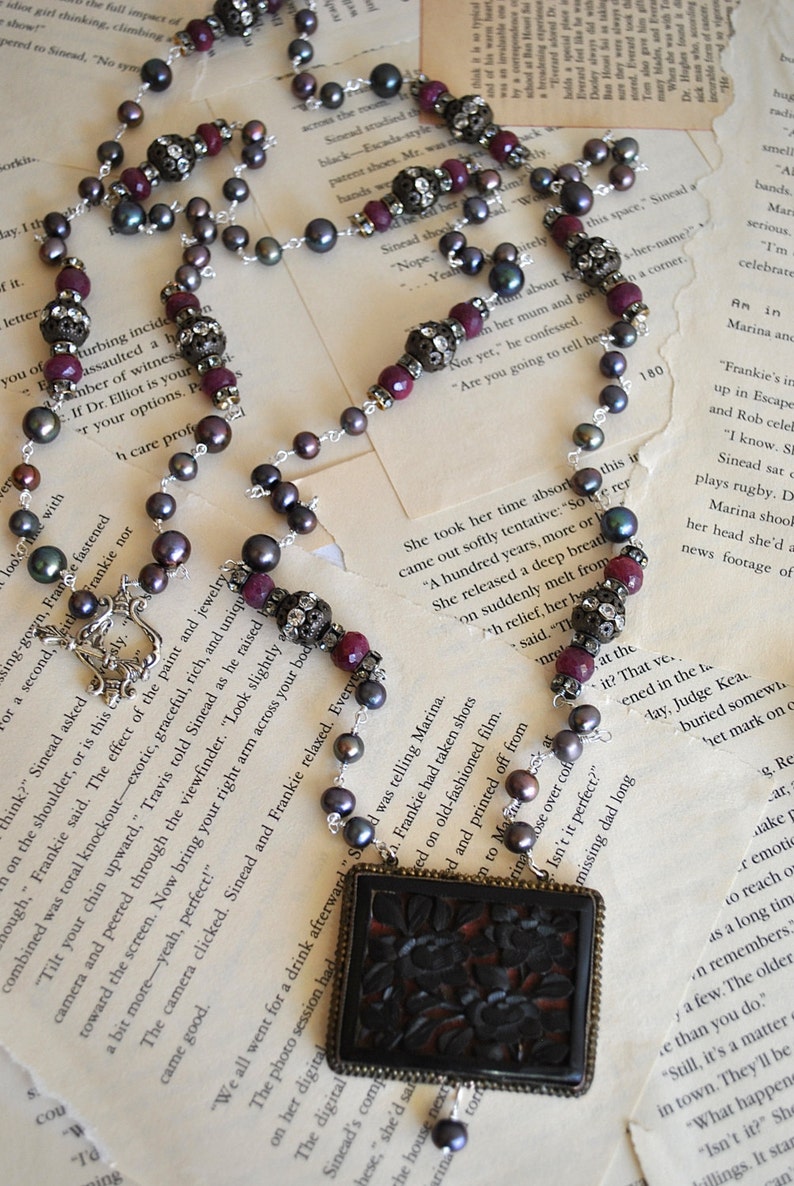 May include: A long necklace featuring dark gray pearls, burgundy beads, and ornate silver accents. The necklace has a rectangular pendant with a dark floral design. A unique piece of jewelry.
