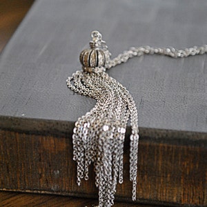 May include: A silver chain necklace with a tassel pendant. The tassel is made of multiple strands of chain and has a crown-shaped top.