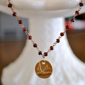 May include: A delicate gold-tone necklace featuring a chain of small, round, deep red beads. A circular pendant with a bird silhouette hangs from the chain. The necklace is displayed on a white pedestal.