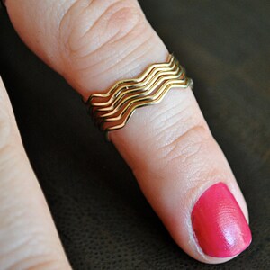 May include: A gold ring with a wavy design, worn on a finger with bright pink nail polish.