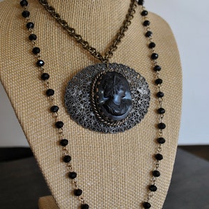 May include: A black cameo pendant necklace with a filigree design and black beads. The necklace is displayed on a brown burlap mannequin.