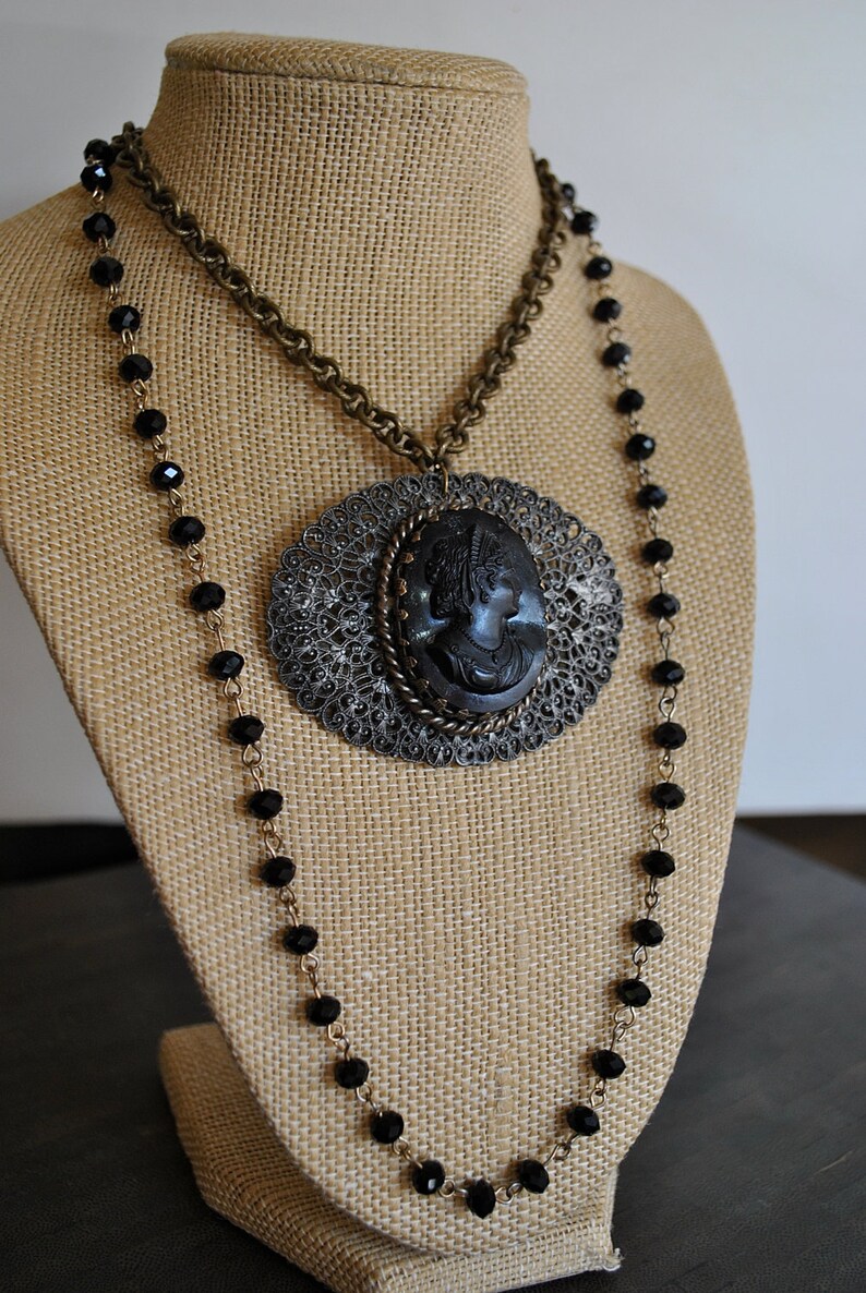 May include: A black cameo pendant necklace with a filigree design and black beads. The necklace is displayed on a brown burlap mannequin.