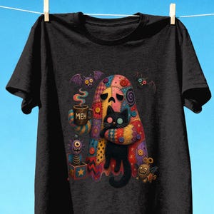 Può includere: Una t-shirt nera a maniche corte con un fantasma colorato e fantasia che tiene in braccio un gatto nero. Il fantasma regge una tazza marrone con la scritta "MEH" stampata sopra. Altri elementi grafici includono piccoli pipistrelli e un giocattolo.