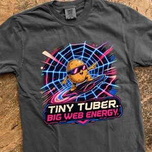 May include: A charcoal gray short-sleeve t-shirt featuring a graphic of a cartoon potato wearing sunglasses and surfing on a neon blue spider web. The text below the graphic reads, TINY TUBER. BIG WEB ENERGY. The shirt has a crew neckline.