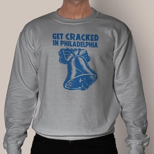 Op de afbeelding: Een grijs sweatshirt met ronde hals en een blauwe grafische print van de Liberty Bell. Boven de bel staat de tekst GET CRACKED IN PHILADELPHIA in een vet, verweerd lettertype. Het sweatshirt heeft lange mouwen en geribbelde boorden.