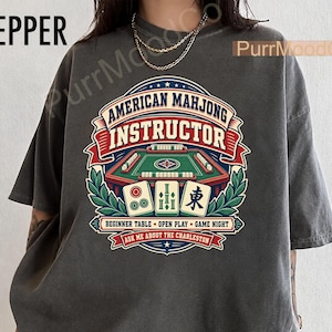 May include: A charcoal gray t-shirt featuring a graphic print with the text American Mahjong Instructor, Beginner Table, Open Play, Game Night, and Ask Me About The Charleston. The design includes a green mahjong table and three game tiles.