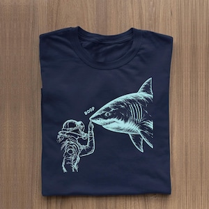 Peut inclure: Un t-shirt bleu marine avec une impression graphique bleu clair représentant un plongeur sous-marin touchant le nez d'un grand requin. Le texte BOOP est imprimé en bleu clair au-dessus du doigt du plongeur en contact avec le museau.