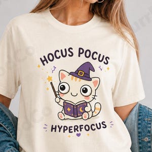 Puede incluir: Camiseta color crema con un gato de dibujos animados que lleva un sombrero de bruja y sostiene una varita y un libro. El texto "Hocus Pocus Hyperfocus" está encima y debajo de la ilustración del gato. La camiseta es de color natural.