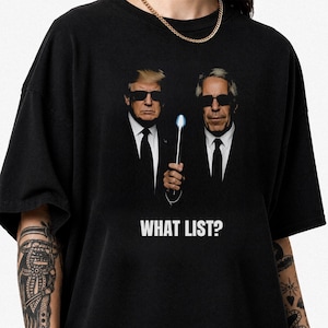 Könnte beinhalten: Ein schwarzes T-Shirt mit einem Grafikdruck von zwei Männern in schwarzen Anzügen und Sonnenbrillen, von denen einer ein leuchtendes Neuralyzer-Gerät hält. Unter den Figuren steht der Text WHAT LIST? in weißen Großbuchstaben gedruckt.