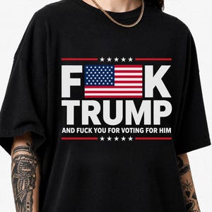 Op de afbeelding: Een zwart t-shirt met korte mouwen en een grafische print met de tekst F*CK TRUMP AND FUCK YOU FOR VOTING FOR HIM. Het ontwerp bevat een vlag van de Verenigde Staten die de letter U vervangt, omlijst door rode lijnen met witte sterren.